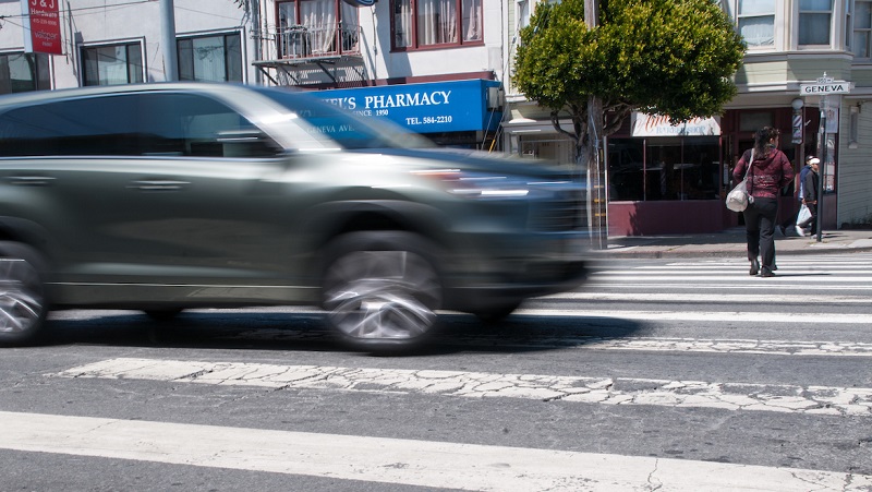 A car appears blurry as it moves through a crosswalk in a San Francisco intersection with pedestrians crossing in the background on Geneva Avenue.