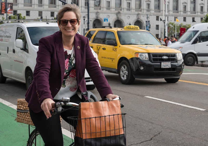 Woman on a bicycle with traffic and Civic Center buildings behind her.