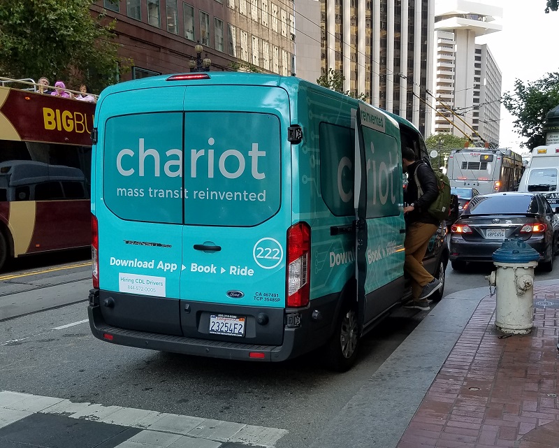 A shuttle van branded with the logo of Chariot, a private transit company, picks up a passenger while stopped in front of a fire hydrant on Market Street.