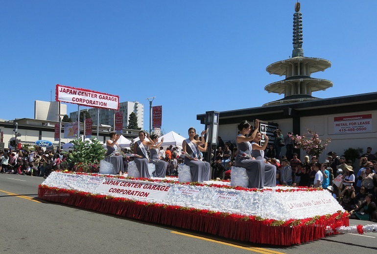 Ladies in gray dresses sit on a red and white float waving to the crowd under a bright blue sky.