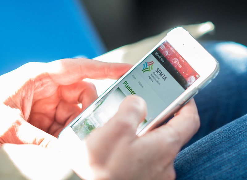 Close-up photo of hands holding a smart phone showing the SFMTA website.