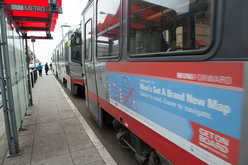 Muni light rail train sits at a transit shelter with a red awning and a Muni Metro logo.