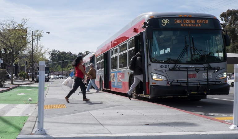 People board a Muni bus at a stop using a new bus boarding island, with a bike lane placed between the island and the sidewalk, on the 9R San Bruno Rapid route.