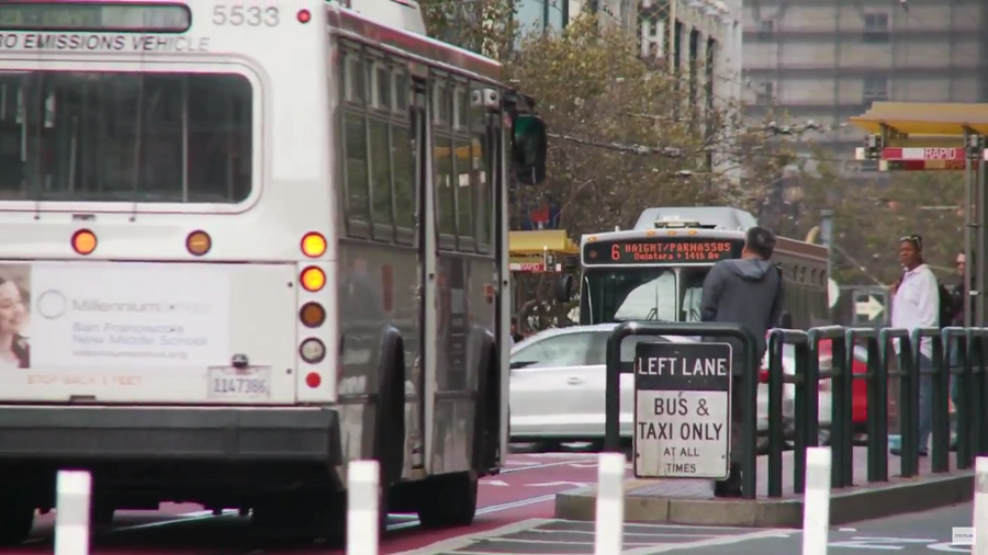 Muni buses on Market Street travel down red-colored transit-only lanes, approaching riders waiting on a boarding island, with a bus shelter labeled, "Muni Rapid."