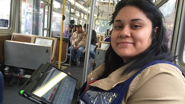 An image of a Muni surveyor in a blue vest and beige coat sitting on a Muni bus holding a black tablet.