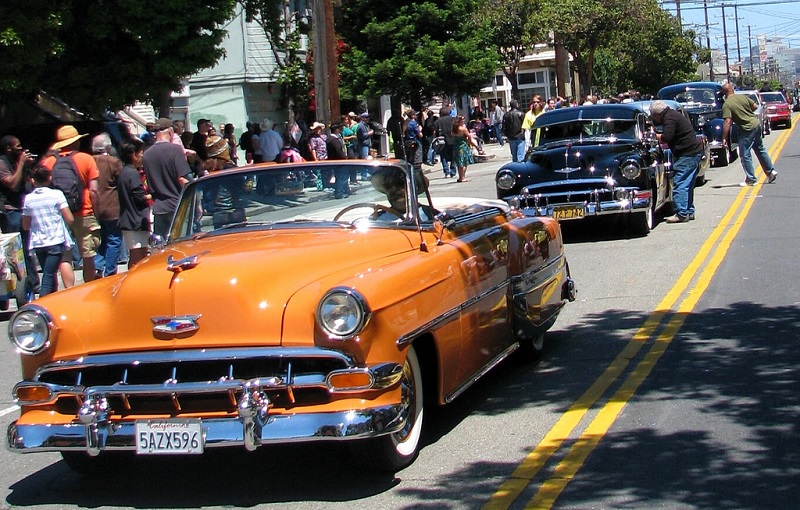 A line of lowrider cars drives down a street with bystanders on the sidewalk in the Mission District on a sunny day.