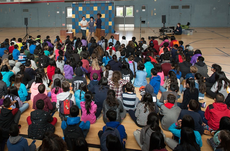 A group of children sit facing three performers on a stage in a gymnasium.