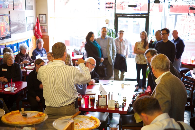 Meeting attendees listen to a speaker in a small pizzeria.