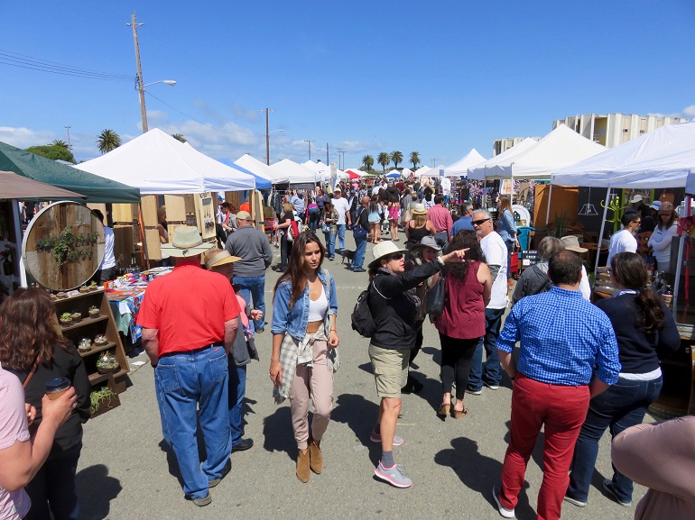 Tent covered vending tables line a narrow walkway on both sides as people look and shop.