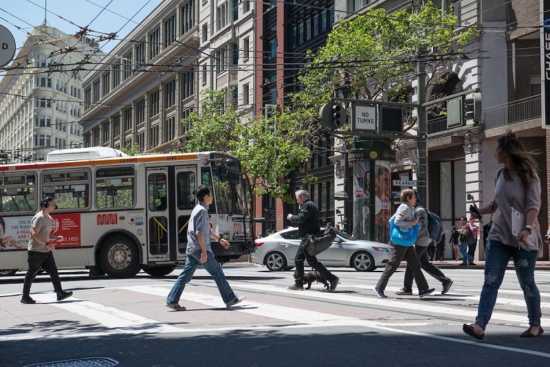 Pedestrians cross a downtown street as a Muni bus and cars cross behind them.