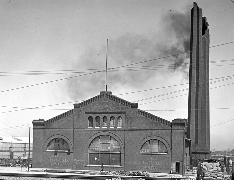 Black and white photo taken in June, 1906 of a large brick building on Market and Valencia Streets. The building is partially damaged with the top of a tall smokestack broken off and light black smoke coming out of it.  Surrounding the building are piles of rubble from destroyed and damaged buildings.