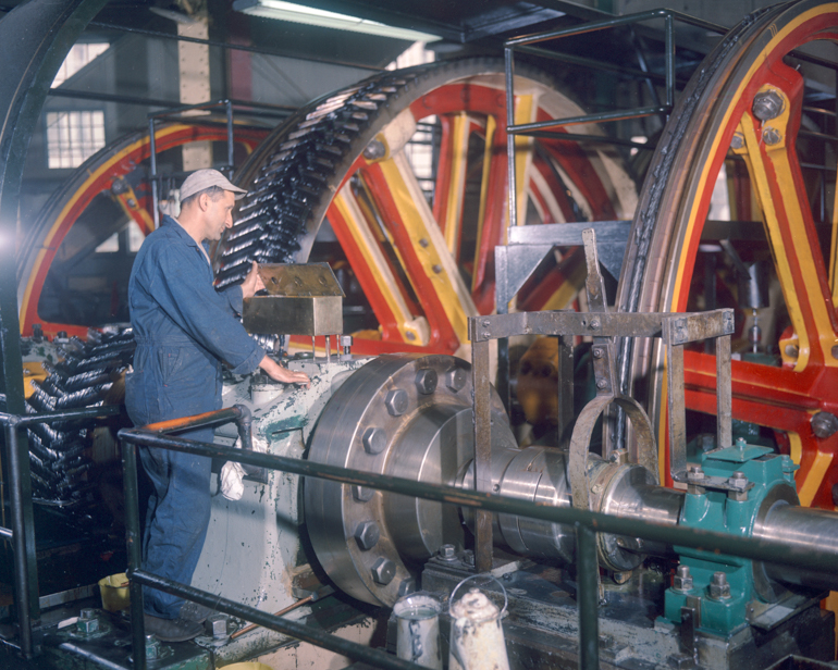 color photo of a man standing next to a huge geared pulley looking into a bronze oil box on top of a large bearing housing.