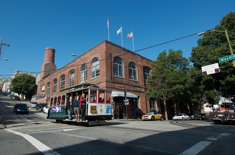 A crowded cable car traveling past the Cable Car Museum and Powerhouse on a sunny day.