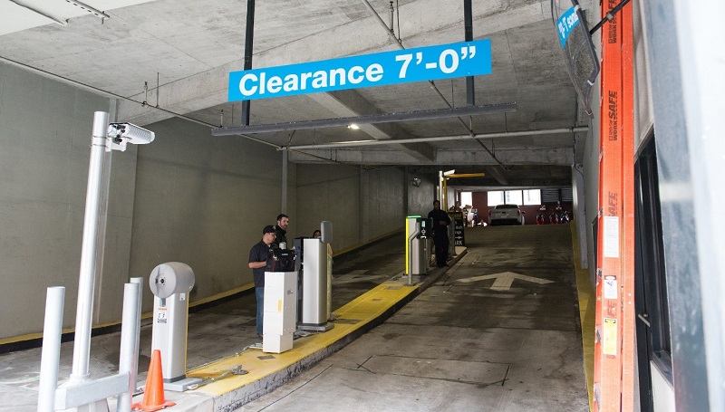 A parking garage entrance ramp with a license plate-detecting camera and several staff members at pay stations alongside the ramp.