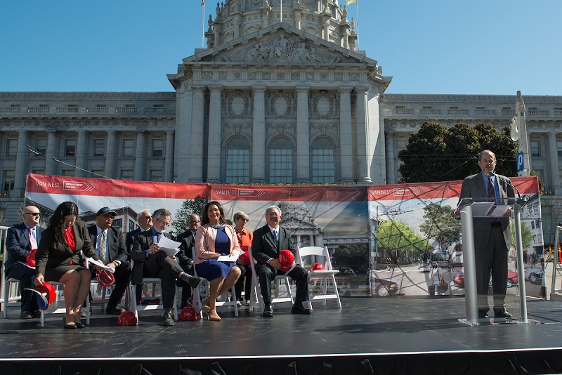 Ed Reiskin speaks at a podium outside San Francisco City Hall. To his right are Mayor Ed Lee, Supervisor London Breed, Supervisor Aaron Peskin, SFCTA Executive Director Tilly Chang, SFMTA Board Chairman Cheryl Brinkman, FTA Regional Administrator Leslie Rogers, Caltrans Director Bijan Sartipi, Public Works Director Mohammed Nuru, and Van Ness BRT CAC Chairman Alex Wilson.