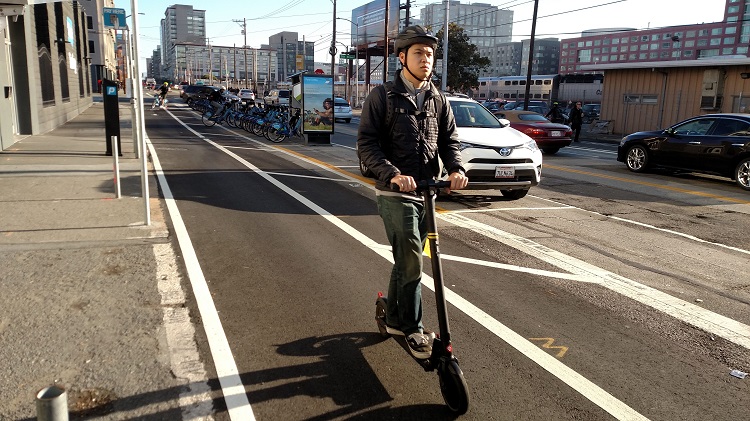 Man with helmet riding scooter in bike lane, with shared bike station in background.
