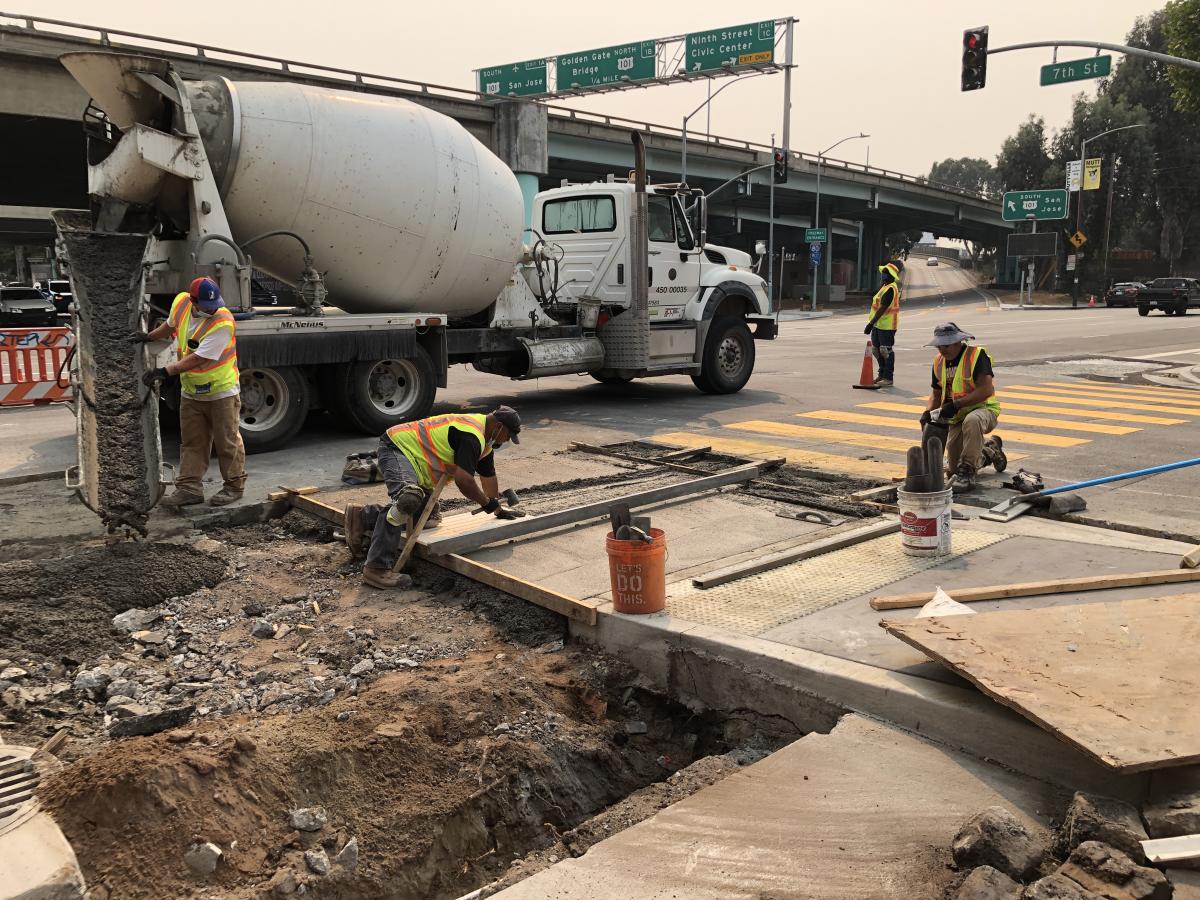 Four people wearing safety vests working on construction of a transit boarding island and a cement mixer in the background