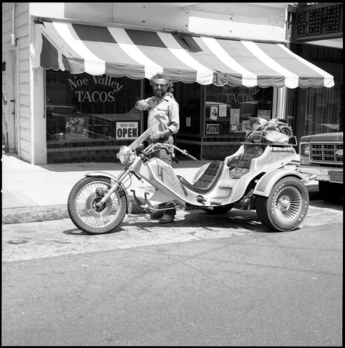 Vintage black and white photograph of a person standing over his motorcycle in front of a restaurant called "Noe Valley Tacos" 