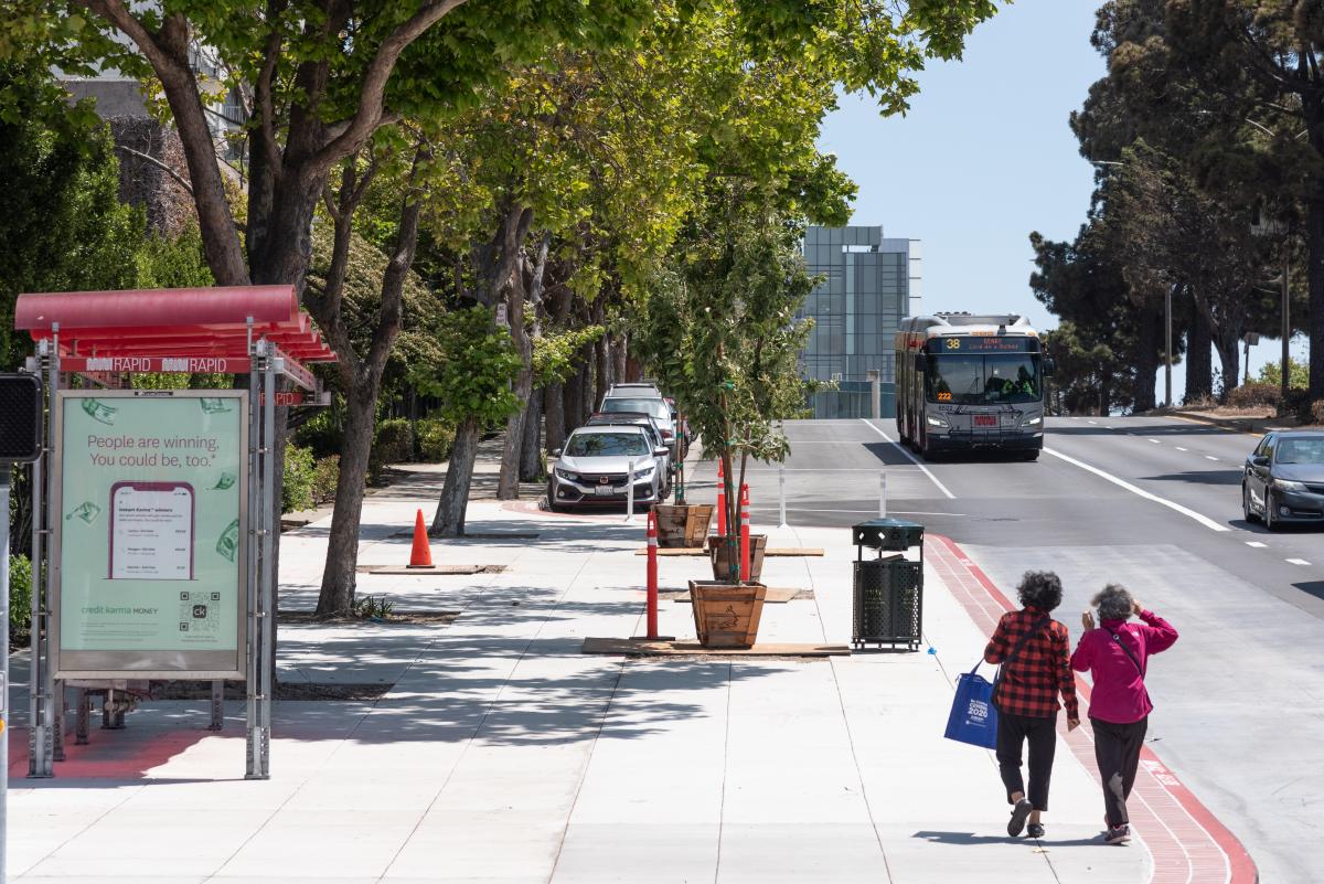 Two people walk on an expanded bus curb amongst vibrant green trees as the 38 Geary arrives