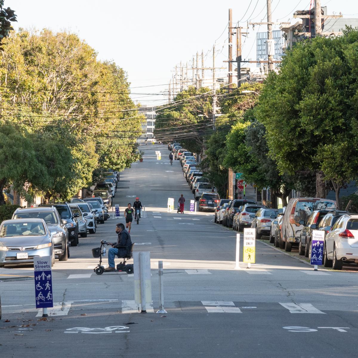 People on Page Slow Street: A man in a motorized scooter crosses the street, and a jogger, a cyclist, and a person pushing a stroller are in the background