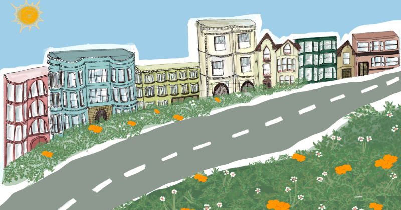 A two-lane road going uphill and flanked by grass and scattered flowers overlooks a row of 2- to 4-story residential buildings of various ages and in different colors.