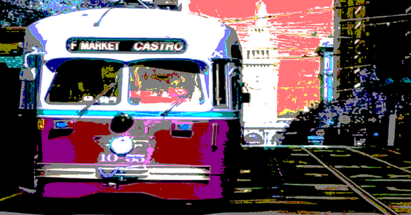 Historic streetcar 1055 with a destination sign of F Market Castro heads toward us on Market Street with the Ferry Building clock tower in the background and trees and office buildings to either side of the street.