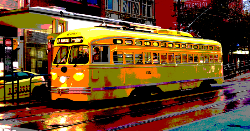 An oblique view of yellow and orange F Market streetcar 1052, with driver and passengers, heading along Market Street toward Fisherman's Wharf. It is at a stop. A Yellow cab passes by. There are buildings in the background, including a theater and a banner for Alonzo King Lines Ballet.