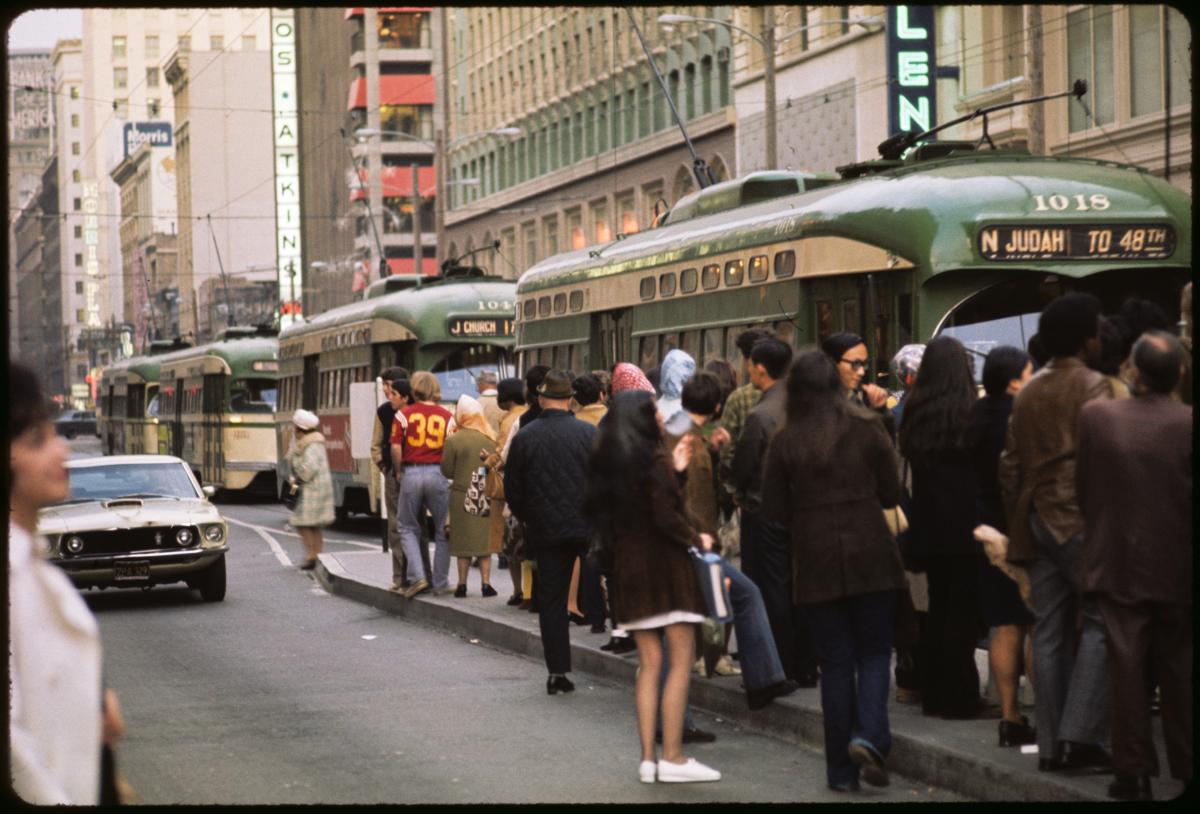 Muni platform overflowing with people as three streetcars approach and a car passes