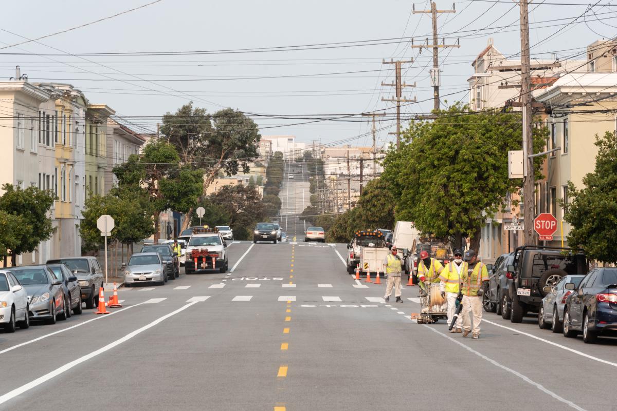 Construction crews striping Anza Street with new bike lanes next to a row of parked cars. The fresh bike lanes are on both sides of the street; there’s a construction truck with cones parked on the left, and cars moving in both directions in the background.