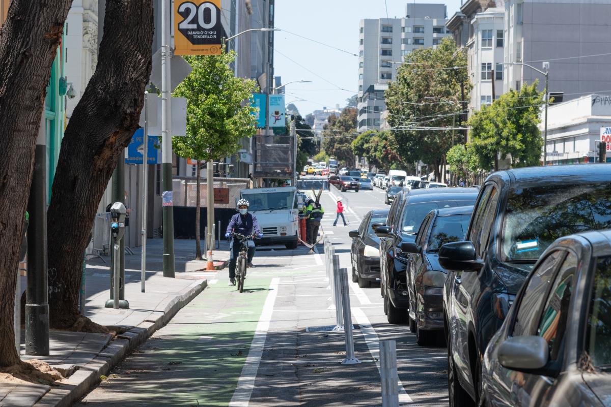 A person rides a bike along the new protected bike lane on Golden Gate Avenue. The lane is painted green, and there are plastic buffer posts between the lane and the parked cars that separate it from traffic. Overhead, a golden-colored sign with 20 on it indicates the new 20 mile per hour speed limits throughout the Tenderloin.