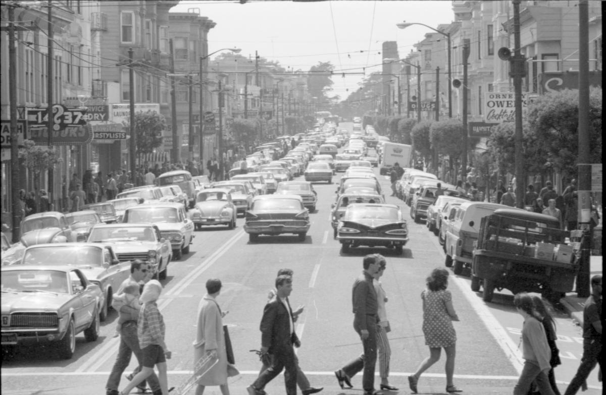 Photo of people crossing street in crosswalk. Street is full of classic cars.