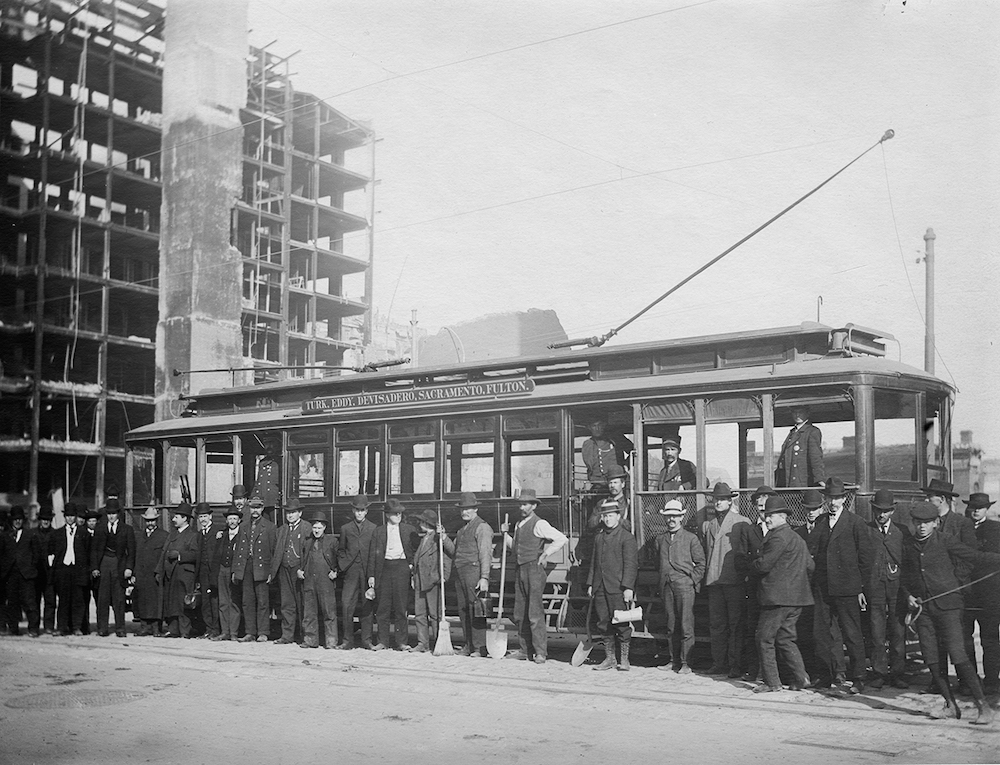 group of people standing along side of streetcar with burned ruins of a building in the background