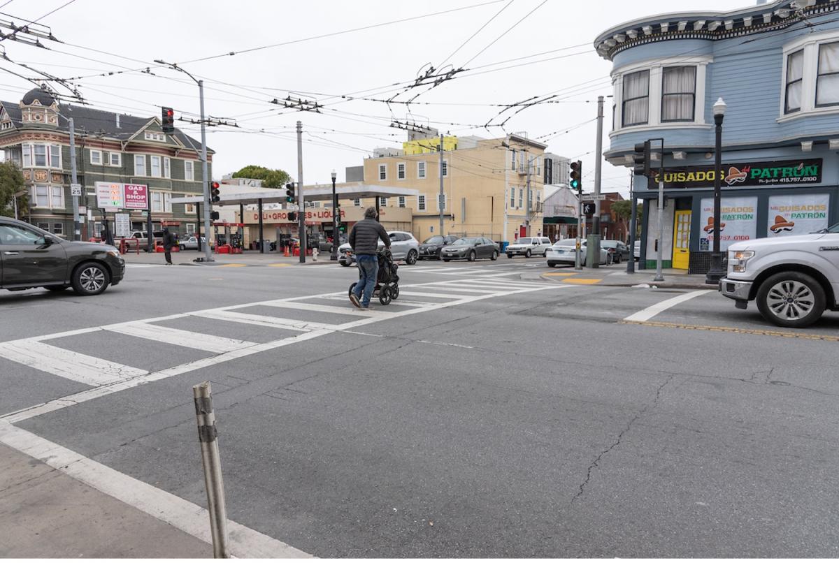 a man pushes a stroller across the street in a crosswalk; a white car is stopped behind a painted white advanced limit line, set back from the intersection