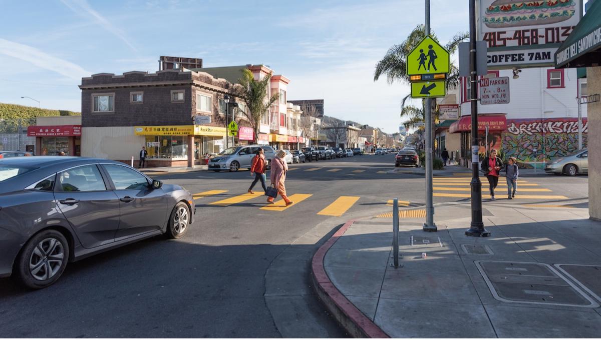 Pedestrians cross a street marked with a continental crosswalk; there is a bright yellow sign at the corner with flashing yellow lights alerting drivers to the people in the intersection