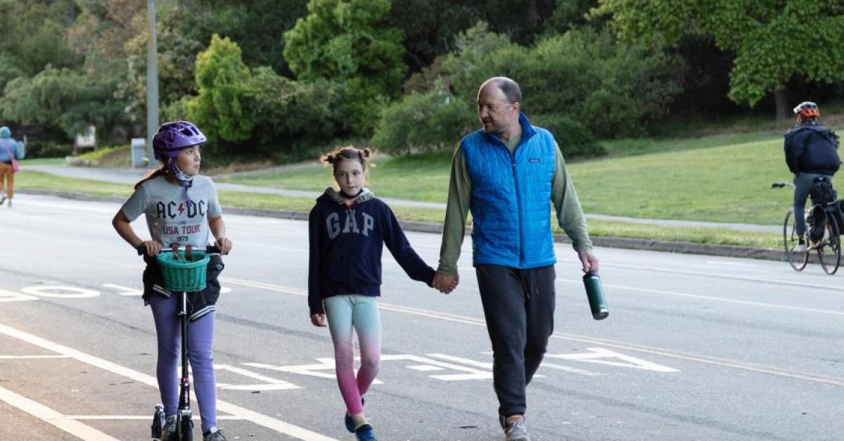 Photo of girl on scooter, man holding child's hand walking, and a person on a bicycle in Golden Gate Park