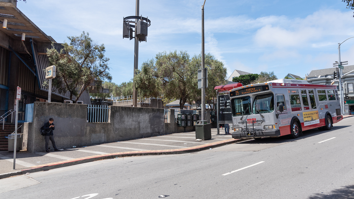 bus in front of stop and sidewalk with BART station in background.