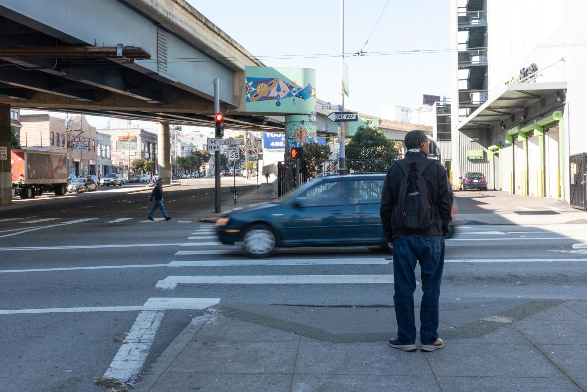 A person is waiting at a intersection for a pedestrian signal. In the background, a vehicle is driving past.