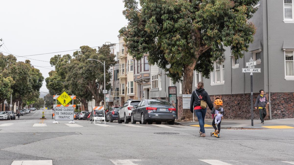 A young kid, accompanied by a woman, rides a scooter through the intersection of Lake and 6th Ave. Slow Streets signs are visible on the street.