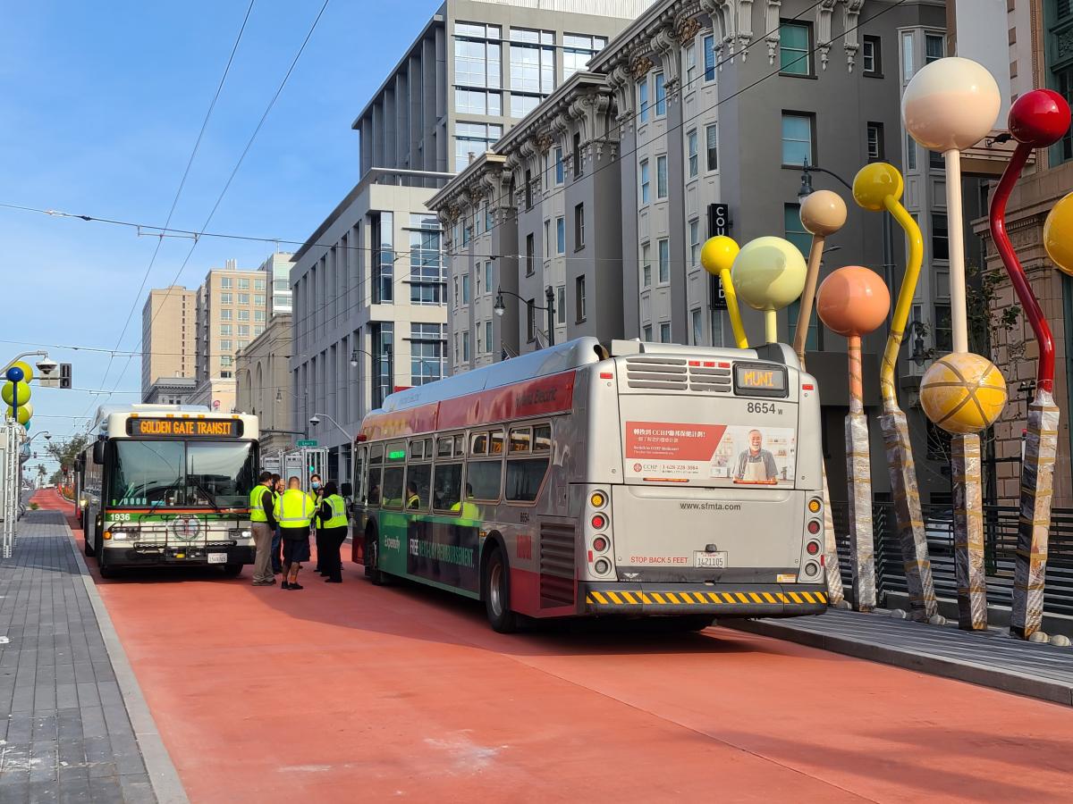 A Golden Gate Transit bus and a Muni bus are parked next to each other in a red transit lane, with staff standing between the two buses. On each side of the platform, public artwork is visible. The public artwork is composed of tall balls in various colors attached to poles, curving in various directions. In the background are mostly housing complexes. 