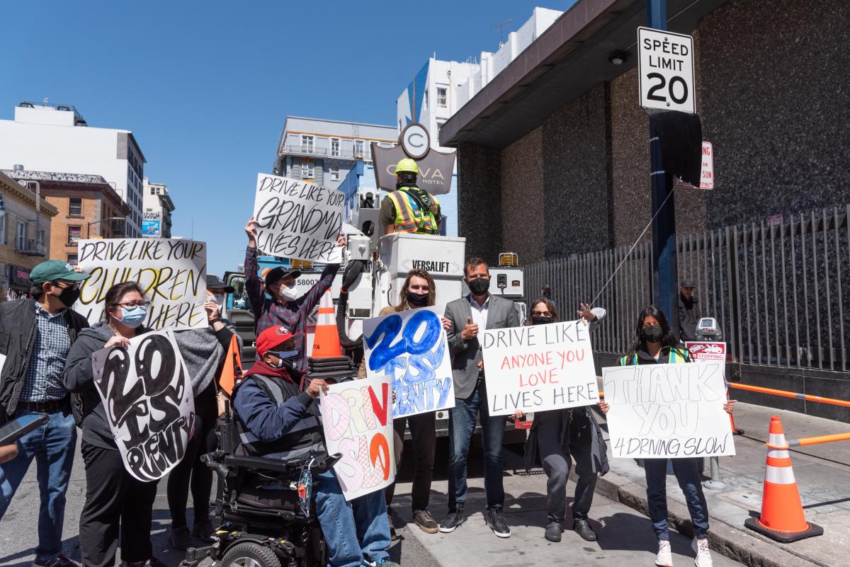 Activists gather in the Tenderloin to unveil new 20 mph signs. They hold signs saying "20 is plenty" and "drive like anyone you love lives here"