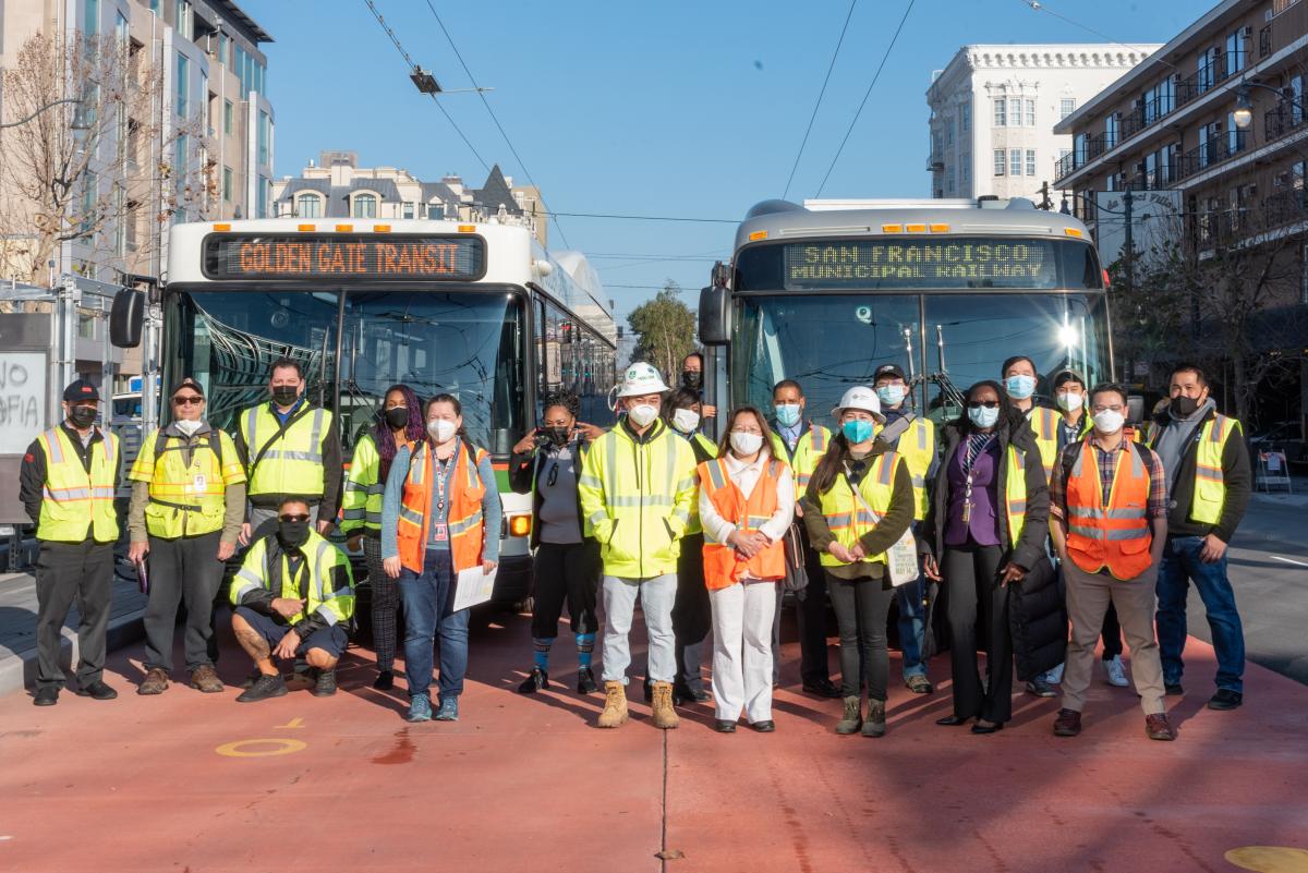 Approximately twenty folks wearing yellow and orange safety vests pose in front of a Golden Gate Transit bus and a Muni bus on the red lanes. Van Ness buildings are visible in the background.    