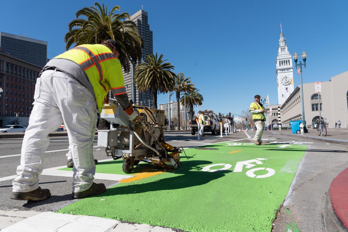 Image of SFMTA paint shop workers striping a green two-way bikeway in front of the Ferry Building on a beautiful sunny day