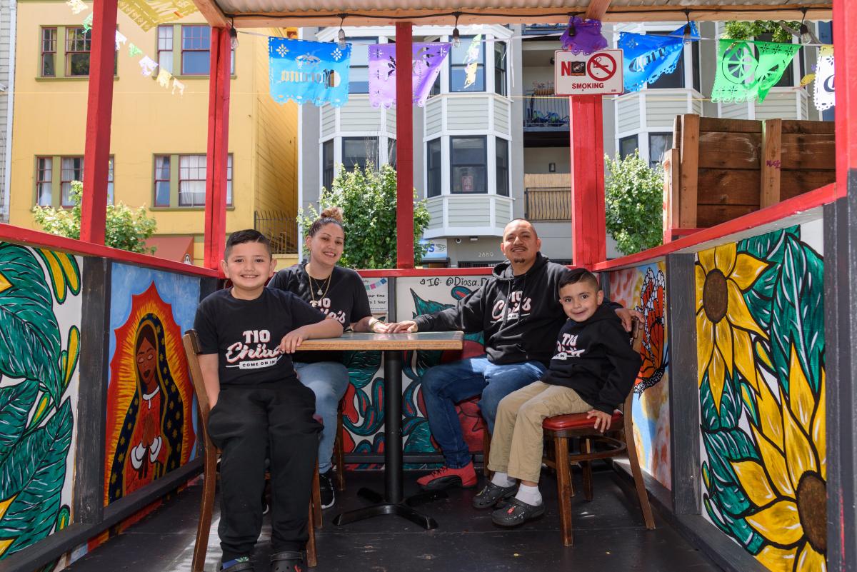 A family shown sitting in a colorful outdoor dining booth. 