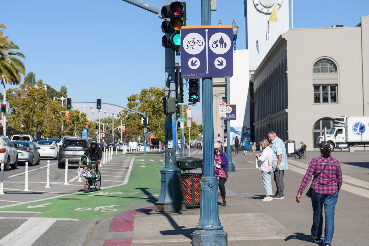 Signs for bicyclist and pedestrians show on a city street filled with cars 