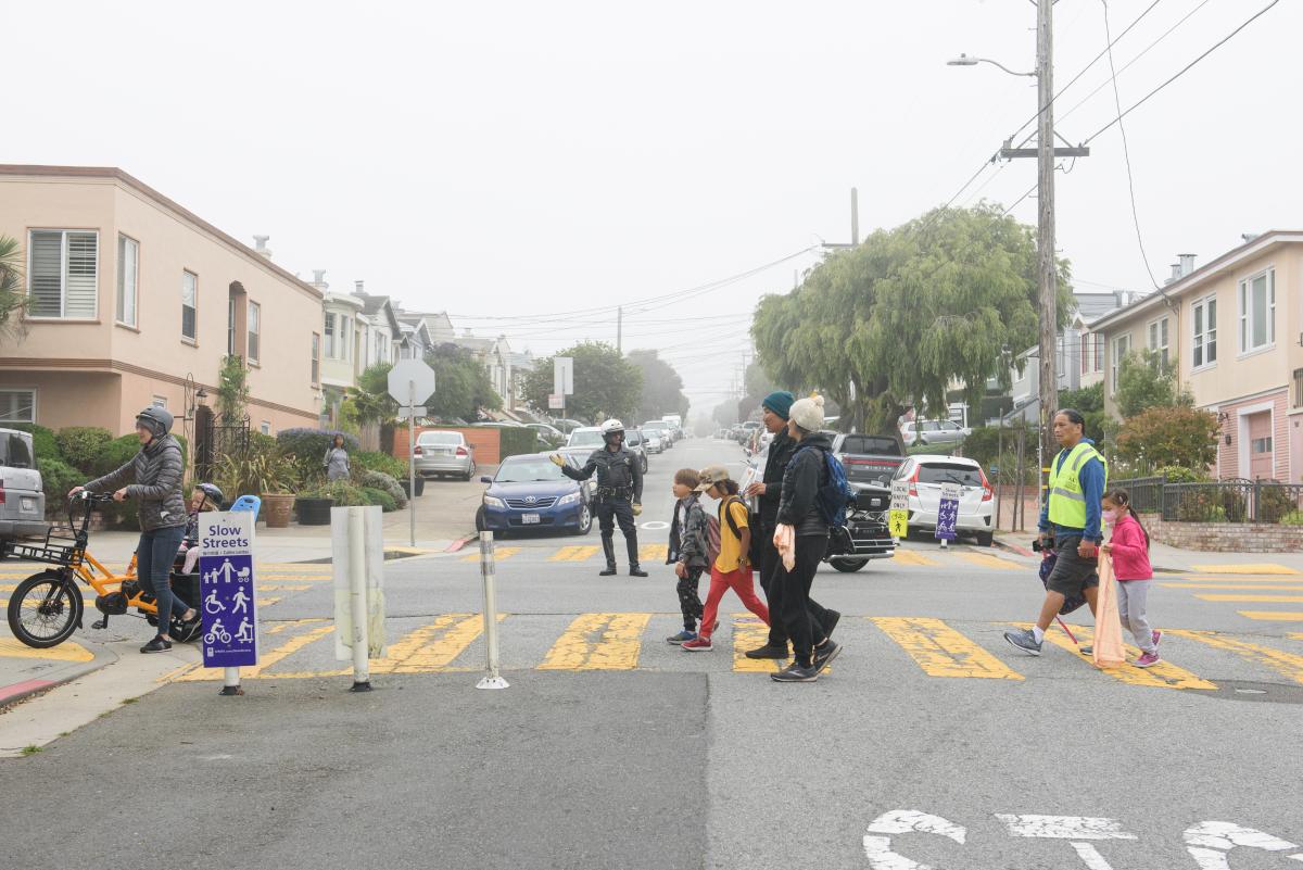 .Children, parents and a bicyclist seen crossing in the crosswalk with a police officer guiding traffic 