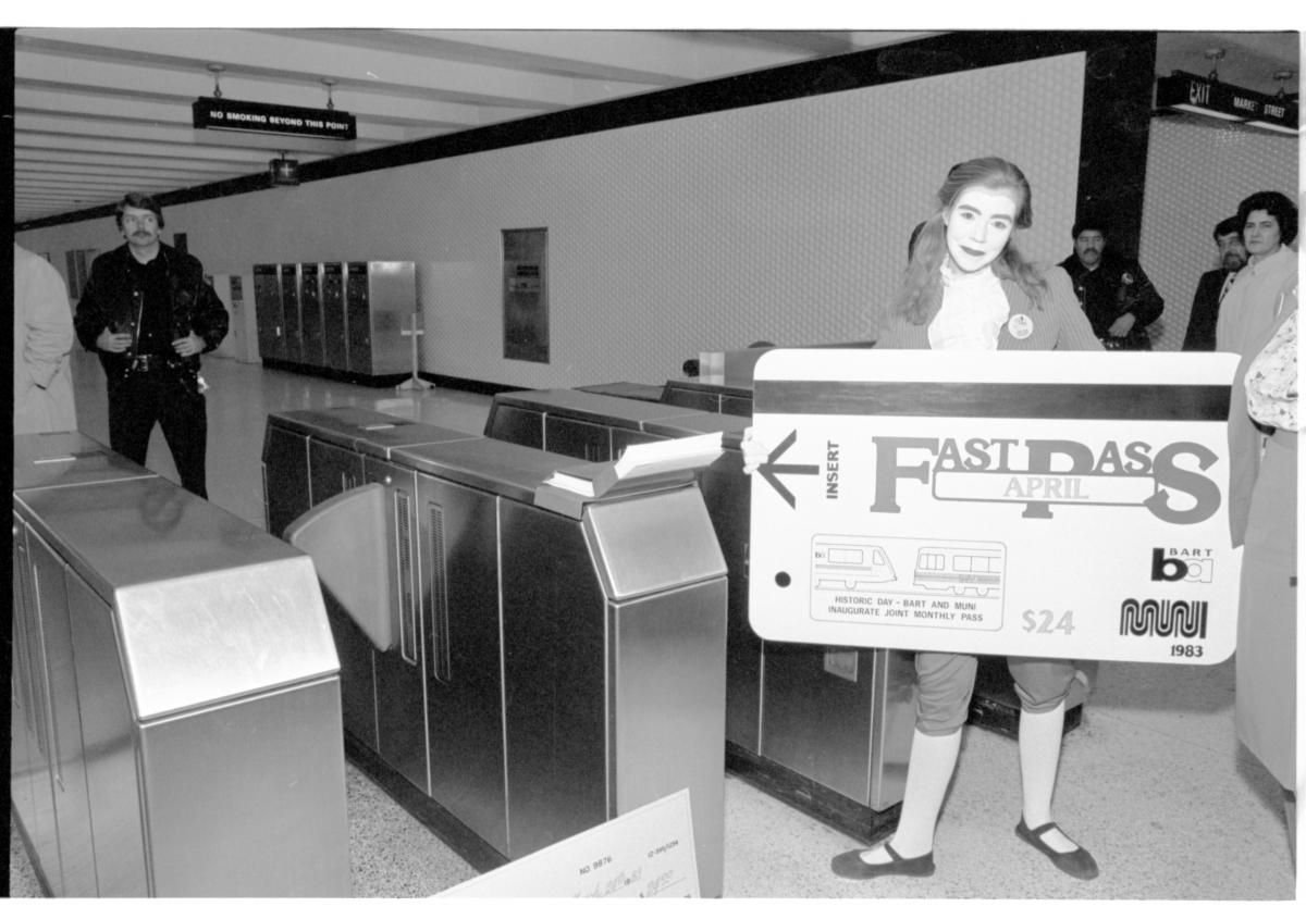 Person dressed as a mime standing next to fare gates holding large replica of Muni Fast Pass transit fare card.