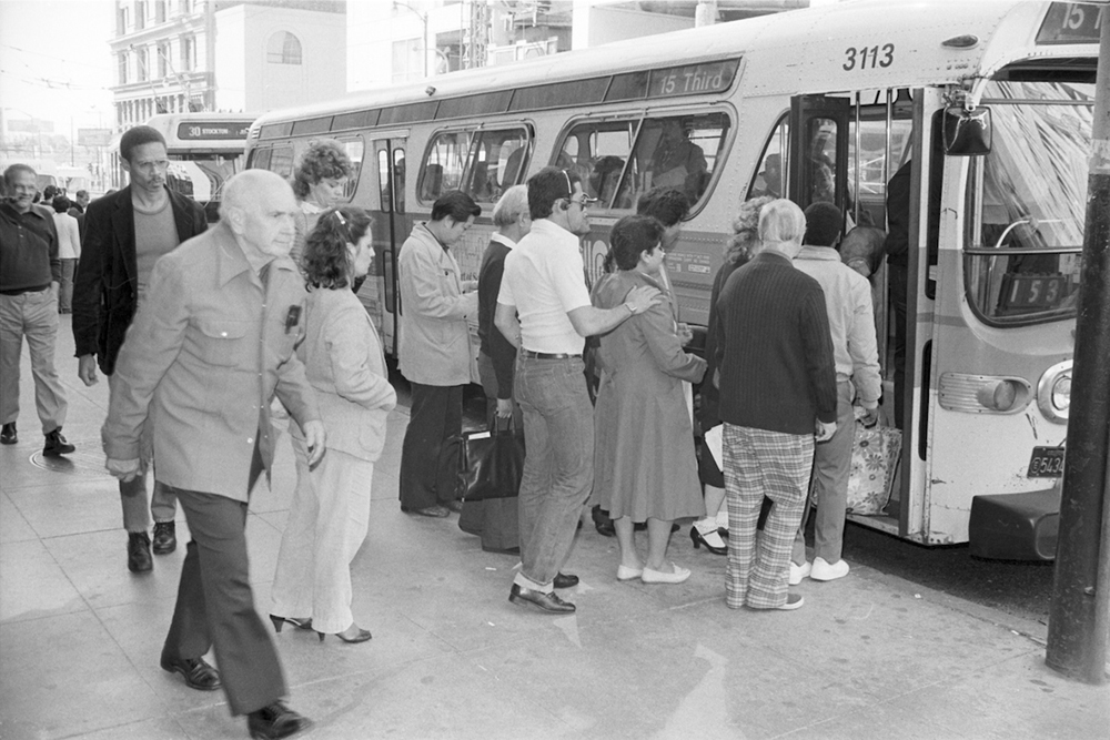 Passengers lining up to board a bus on a busy city street with buildings in the background 