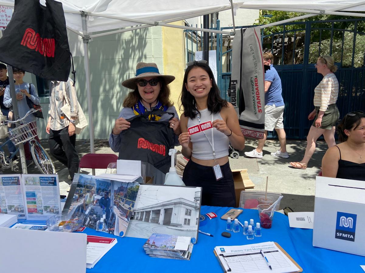 Two people standing behind a table are seen smiling at the camera. They hold Muni-labeled items.