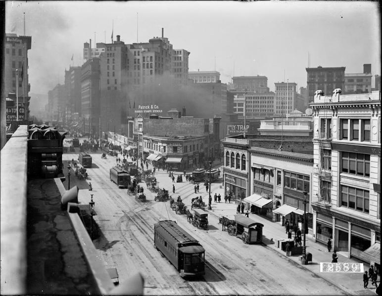 Black and white overhead view of street with horse-drawn and electric streetcars, horse drawn wagons, pedestrians, and buildings. Business signage includes Patrick and Company and Fly Trap.