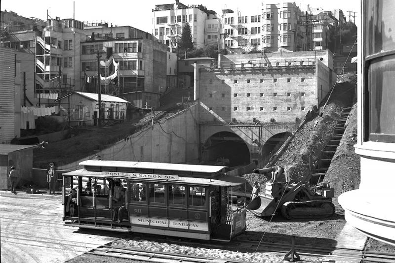 cable car in foreground with excavation, bulldozer, and tunnel entrance in background. car is branded "Municipal Railway" and signed "Powell and Mason streets".